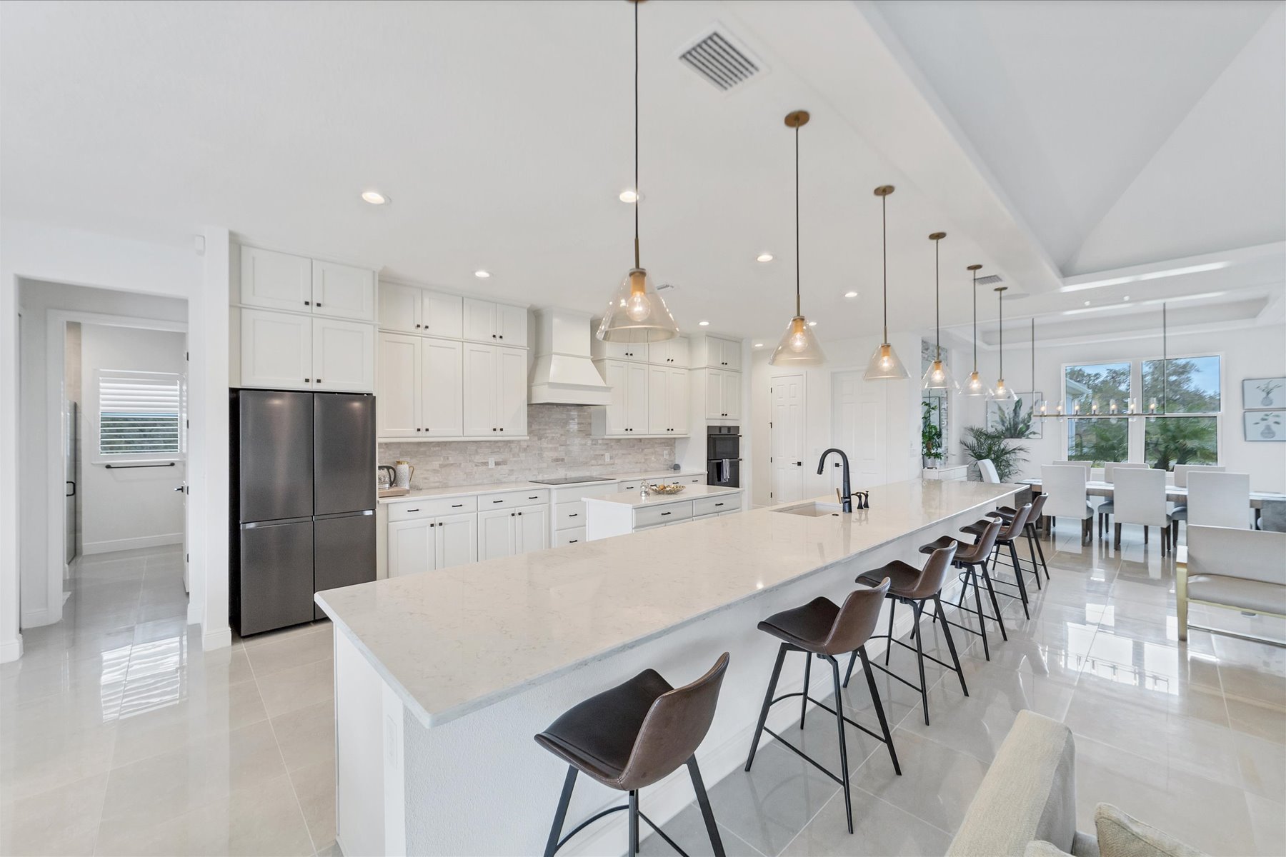 Open white kitchen with a long quartz island, brown leather stools and glass pendant lights